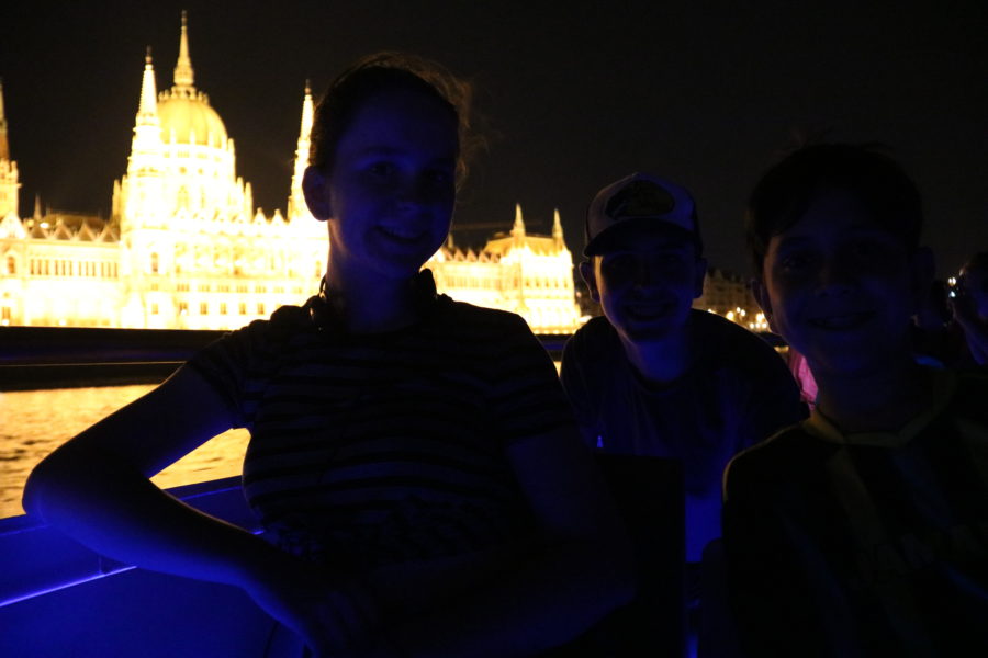 image of my kids in Budapest on top floor boat with Parliament glowing in background at night