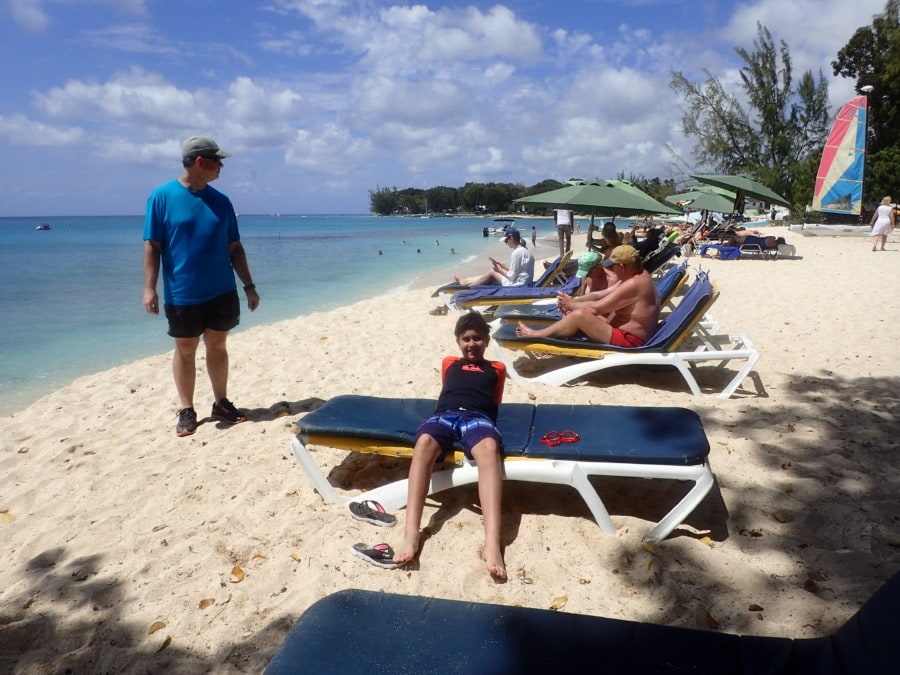 John standing beside Caiden who is sitting on blue lounge chair on beach in Barbados