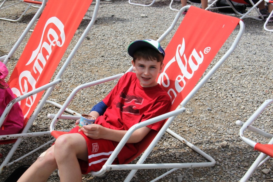 kid with baseball hat and red tshirt with blue bandage on right arm sitting on red lawn chair with Cola written on it