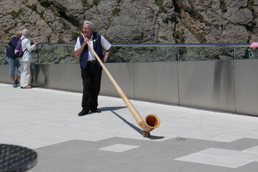 man with black pants and blue vest over white shirt holding a very long light beige alphorn at top of Mount Pilatus