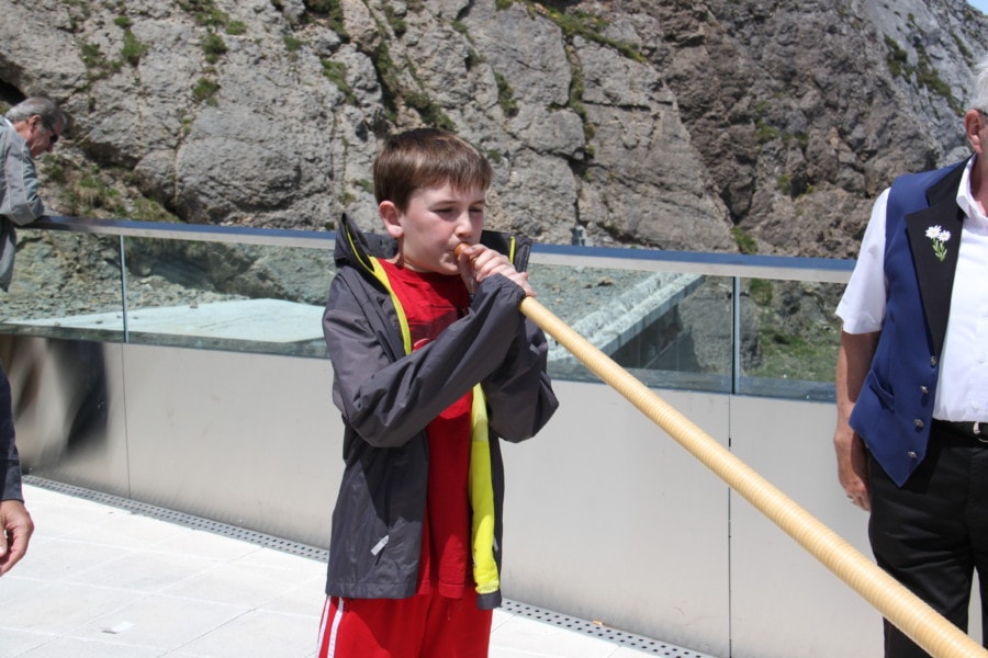 Lucas wearing red shirt and grey jacket blowing into alphorn
