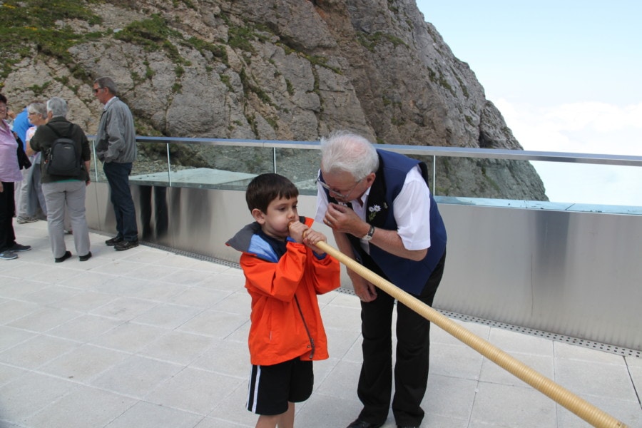 Caiden with orange jacket blowing alphorn while man teaches him how to blow