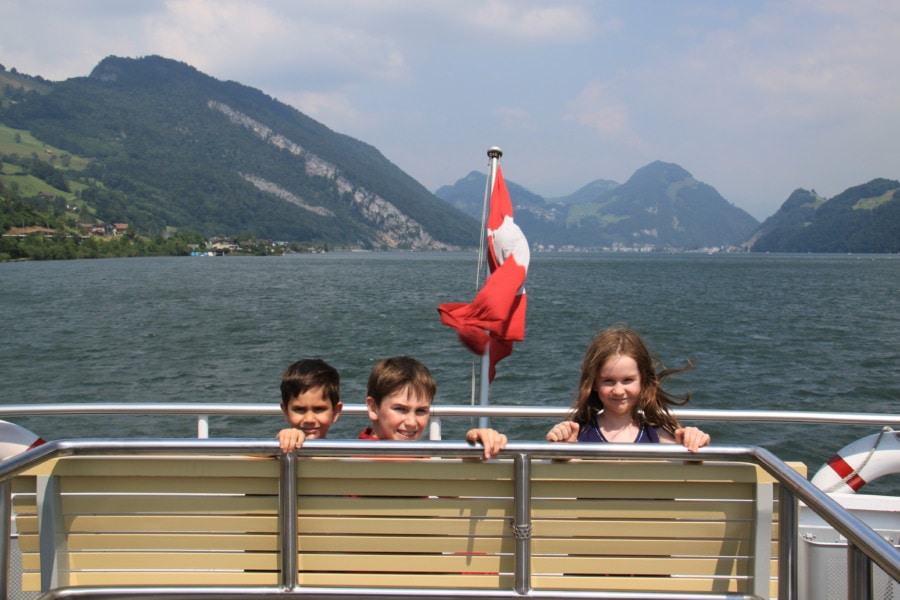 three kids sitting on front bench with Swiss flag between them turned around with mountains behind them