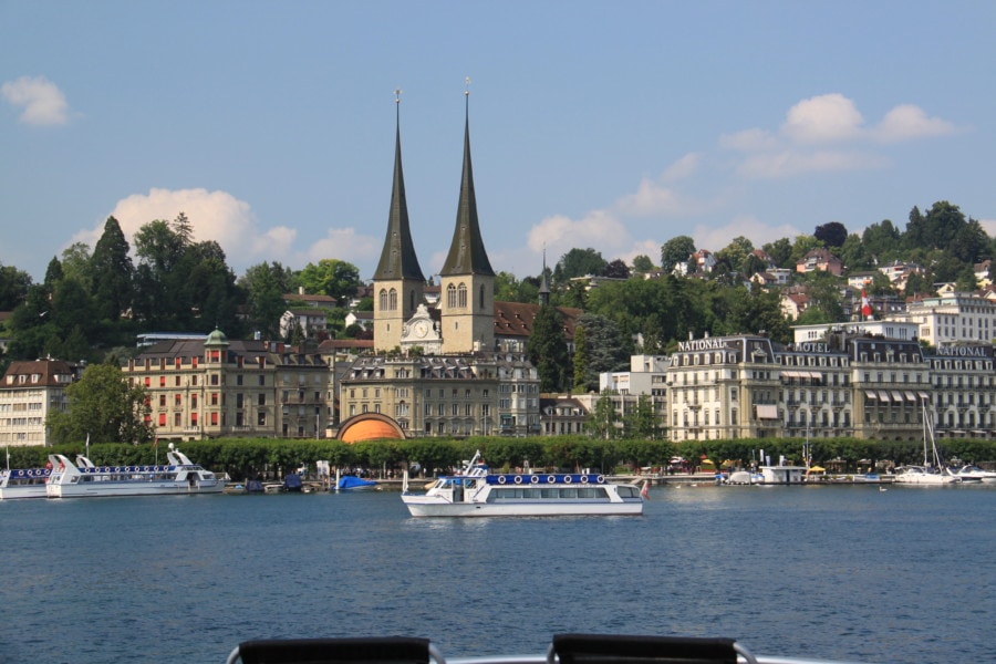 view of Lucerne from the water seeing another ferry, dock, beautiful double steeple church, and large square buildings