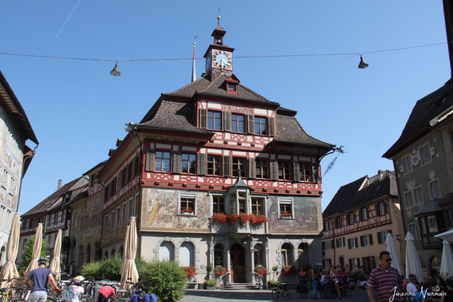 town hall in Stein am Rhein is four stories with clock tower on top and each layer is painted differently with grey on bottom, frescos and scenery in middle and white with red trim on third and fourth level