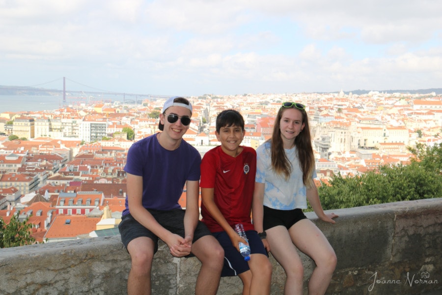 My three kids sitting on a short wall with Lisbon orange roof tops and Tagus River behind 