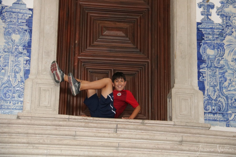 Caiden in red shirt having fun with feet in air in front of brown wooden door at top of stairs super fun thing to do in alfama with kids