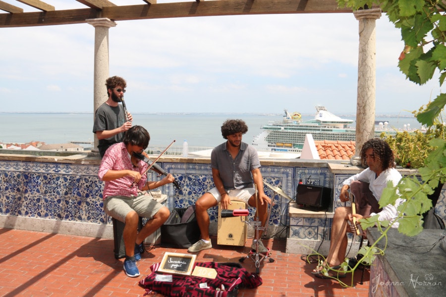 four musicians playing fiddle, clarinet, drum and guitar on edge of terrace with river and cruise ship behind