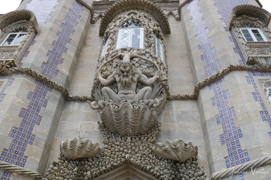 arch leading to terrace is white and blue tiled building with  sea monster holding up bowed window and decorative shells