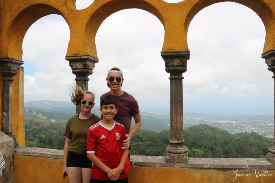 three kids with hair blowing in front of yellow arches overlooking Sintra village on daytrip from Lisbon