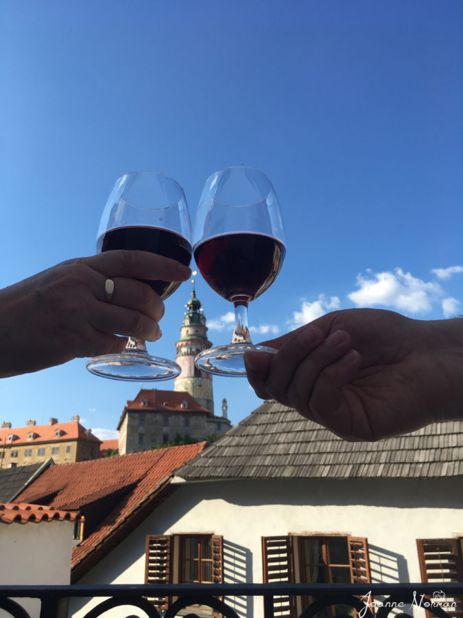 us holding two wine glass which frame krumlov castle tower in distance