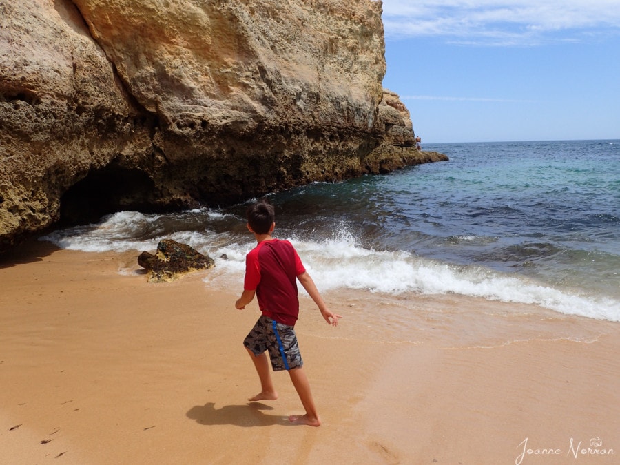 Caiden in red shirt running from waves next to rock cliff at Praia de Vale Centeanes