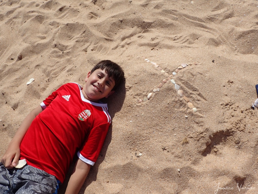 Boy in red lying on sand with shells creating an X to the right side 