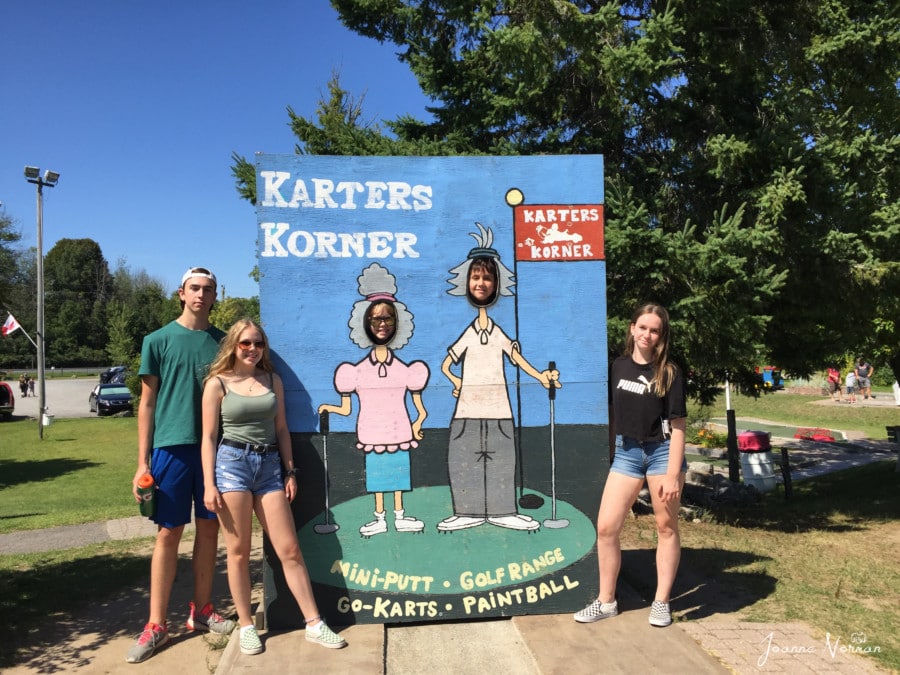 Karters Korners sign with holes for faces and five teens standing near 