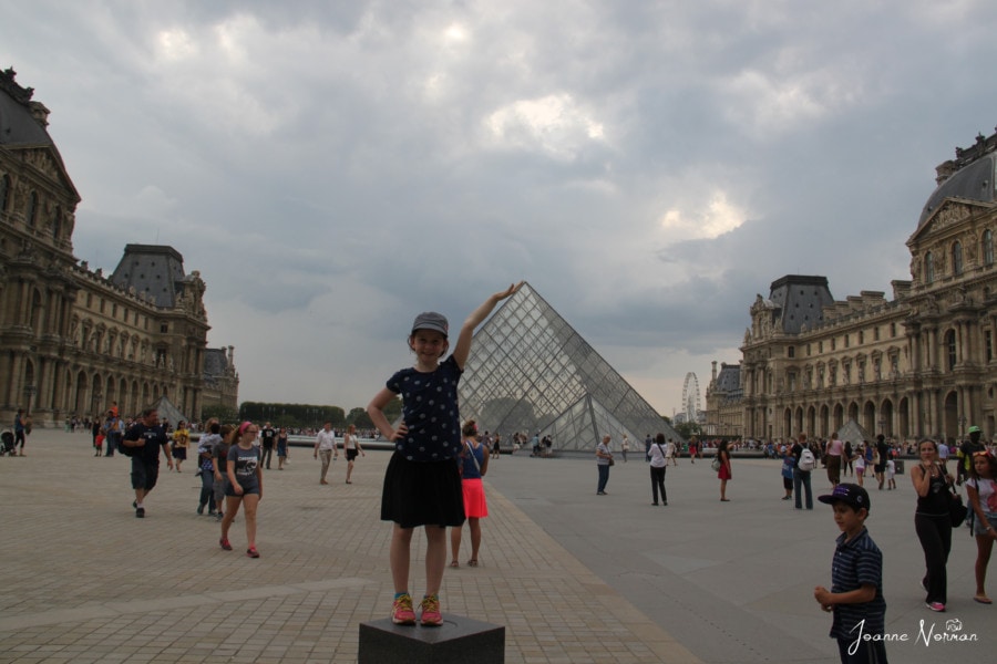 girl standing on block with hand in air so it looks like she is holding the Louvre pyramid Paris holidays with kids