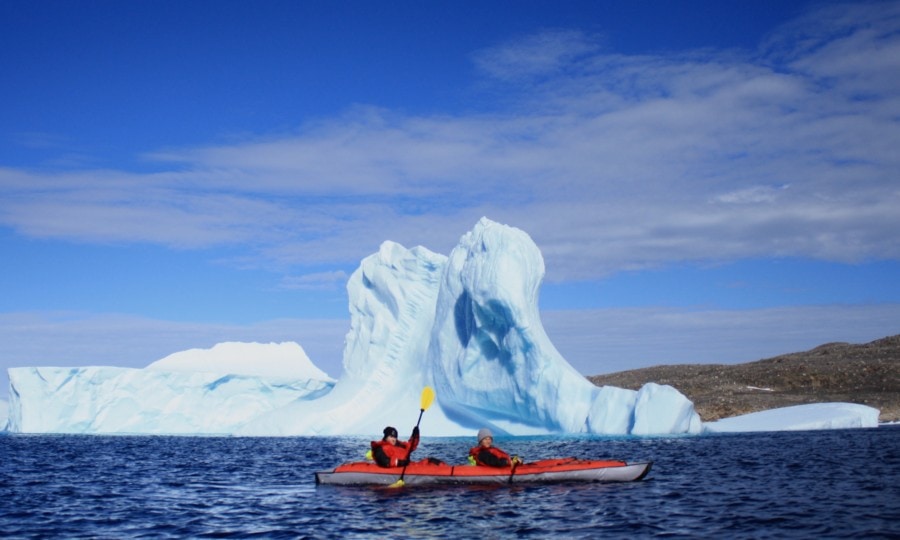 two people in red kayak paddling by tall white ice burg during autumn in arctic